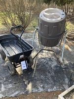 Photo showing the Suncast compost bin on metal stand next to the black garden wagon on concrete pavement beside bushes.