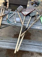 Garden tools on a wooden picnic table featuring wooden handle rakes, post hole digger, mallet axe, bulb planter, shovels, and snippers.