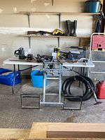 Wide view showing work table with power tools including band saw, heat gun, electric drill, propane tanks, hose, boots and gas can below shelves with hand saws and levels