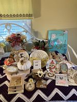 Wide view of 21 handcrafted home decor pieces arranged on a table, including plates, floral arrangements, artwork, and seasonal accents in various colors and designs.