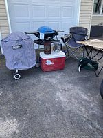 Photo showing two Weber grills, one covered kettle charcoal grill and one blue portable propane grill, along with a red and white cooler, two charcoal chimney starters, a gray foldable camping chair, and a wooden slat folding camp table.