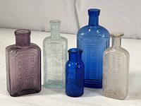 All five antique pharmacy bottles showing variety in colors: cobalt blue, amethyst, green-tinted, and clear glass, and various sizes from 4 to 7 inches tall.