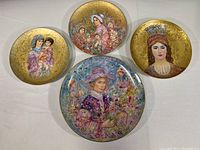 Four Edna Hibel limited edition collector plates arranged on a white surface showing various portrait scenes of women and children with gold backgrounds.