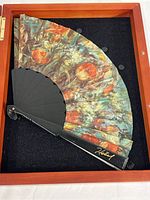 Fabric expanding hand fan with floral print displayed folded inside a wood shadow box case with black felt background. The fan has a black frame and is partially open.