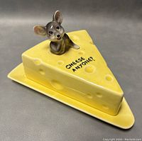 Two angled views of a yellow ceramic cheese dish in the shape of a Swiss cheese wedge with a mouse figurine and text 'CHEESE ANYONE?'. Mounted on a matching base plate.