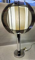 Photo of the lamp fully assembled and lit, showing spherical acrylic panels around fabric shade on chrome pedestal base.