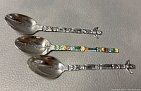 Photo showing three sterling silver collector's spoons laid side by side on a textured leather surface, highlights totem pole designs and 'ALASKA' engraved on bowls.