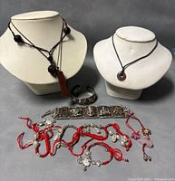 Overall collection including two ivory-colored necklace displays showing two brown cord necklaces with carved stone pendants, silver-tone metal cuff and link bracelets, and several red string charm bracelets with pendants.