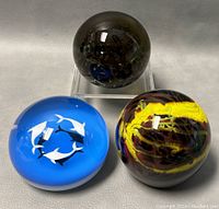 Three Baccarat 3-inch blown glass paperweights shown, including dark smoke bubble design, blue dolphin figure design, and abstract multicolor swirl design.
