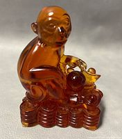 Front angle view of the amber-colored glass monkey sculpture sitting on a base of stacked coins, showing the monkey holding a ball and a gold ingot.