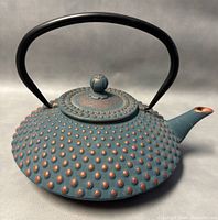 Front angled view of teal blue-green cast iron teapot with rust-colored raised dots and black handle.