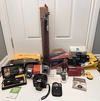 Overview photo showing multiple vintage cameras including Konica Auto S3, Vivitar 600 flash in box, Argus camera, Minolta camera, original boxes, tripod, film rolls, camera cases, and accessories.