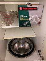 Toastmaster hand mixer in original box along with glass measuring cup and nested stainless steel bowls on a white shelf.
