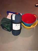 Camping lot items on floor with green soft cooler bag, Woodstock sleeping bag in navy blue storage bag, blue and white hard plastic cooler, and three colorful plastic buckets (red, yellow, blue)