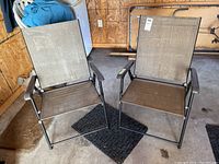 Two folding outdoor chairs with metal frames, plastic arms, mesh fabric seats and backs placed side by side on a concrete garage floor with walls of unfinished wood paneling.