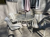 Four outdoor armchairs with metal frames and striped synthetic fabric in beige and taupe tones around glass outdoor table. Deck and fence background.