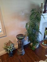 Tall leafy faux plant in white planter, black weathered classical column pedestal, and shorter black urn-shaped planter with spherical plant arrangement next to a wall