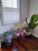 Wide shot showing all three faux plants including blue and white ceramic planter on wooden stand, colorful flower bouquet, and tall plant with large green leaves.