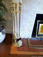 Full view of brass fireplace tool set with stand showing the shovel, broom, poker, and tongs leaning against the stand. Set positioned on brick hearth in front of white painted brick fireplace.