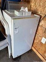 Fridge shown at an angle with a dent visible on the front door left side and some dirt marks near the base. Multiple plastic chairs located beside the fridge, and a wooden panel wall behind it.