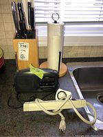 Photo showing wooden knife block with knives and scissors, paper towel holder with partial paper towel roll, GE portable radio, and white power extension cord on kitchen counter.