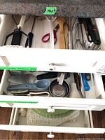 Open kitchen drawer containing measuring cups, ladle, tongs, and other kitchen utensils organized in a white tray.