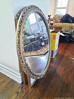 Full view of the oval mirror showing reflective glass and decorative gold frame standing on hardwood floor.