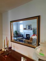 Front view of large rectangular framed bevelled wall mirror hanging on white wall, reflecting living room with fireplace and furniture.