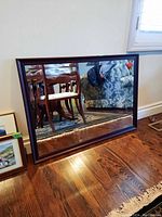 Mirror leaning against a white wall on a hardwood floor, showing reflection of furniture and room interior.