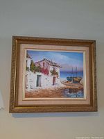 Framed canvas painting depicting two whitewashed buildings with greenery, near a beach with boats, bright blue sky and mountains in background, signed lower right.