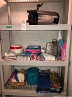 Shelving displaying Westbend coffee maker on top shelf and various party supplies including plates, cups, napkins, utensils on lower shelves.