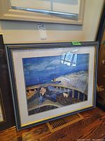 Framed print showing a person lying on a curved wooden boat structure with ocean and shoreline background under a cloudy sky, in a dark wood frame with gold trim.