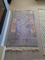 Full view of wool rug laid flat on carpeted floor showing floral design with muted grey-purple background and beige borders with floral and geometric patterns. Fringes visible on short edges.