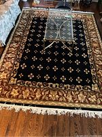 Top-down view of the full rug on a wooden floor with a glass coffee table placed partially on it, showing the floral border, central pattern, and fringe clearly.