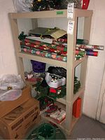 Front view of the shelving unit with Christmas wrapping paper and decorations on shelves. Cardboard box and other storage items nearby.