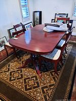View of the wooden dining table with five chairs around it, tablecloths and protector on top, placed on patterned carpet.