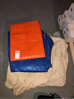 Photo showing three tarps and drop cloths stacked on floor: orange synthetic tarp, blue synthetic tarp, and beige paint drop cloth.