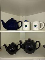 Four tea pots and two white mugs and a creamer and sugar bowl on shelves.