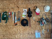 Wide shot of tools and accessories hanging on a garage wall over oriented strand board, showing multiple saws, a green Craftsman electric trimmer, hose sprayer and hardware items.
