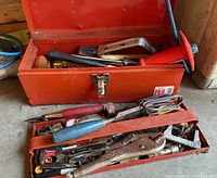 Open red metal toolbox with tray showing various hand tools inside including chisels, screwdrivers, hammer, pliers, tape, and clamps.