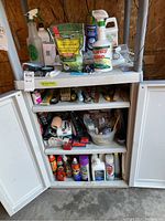 Wide shot showing white cabinet with open doors revealing shelves stocked with various garden chemicals, spray bottles, lubricants, and hand tools.