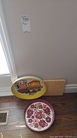 Photo showing two round metal trays with different colorful prints leaning against wall on wooden floor.
