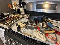 Wide shot of trays with assortment of kitchen utensils including graters, spoons,whisks, flippers, candy thermometer and Cuisinart mixer beater on stove.