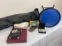 Photo showing blue foldable stool, grey camp chair bag, folded handkerchiefs, box of hair curlers, antique razors, Scrabble game, decks of cards, and necklace box arranged on white surface
