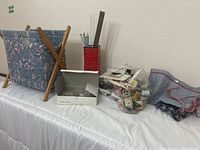 Photo of wooden knitting basket, box of needles, red container holding knitting needles and ruler, plastic bag of threads and pins on table
