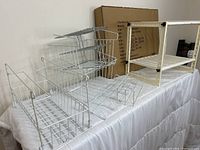 Three assorted white metal wire shelving units on a white surface - two with tiered basket designs and one with a frame and slatted wire shelf, all suitable for under sink or closet organization.