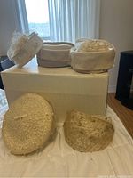 Five beige vintage women's hats arranged on and in front of a beige box on a white surface, showing variety of styles including pillbox and wide-brim with mesh and woven textures.
