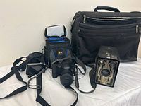 Overall lot showing three cameras and three camera bags against a white wall and floor surface.