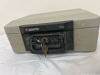 Front view of closed Sentry 1100 portable safe showing beige textured case, black front lock handle, and included keys.