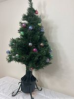 Full view of artificial Christmas tree with LED-lit branches and colorful Christmas balls on branches, standing on a table.